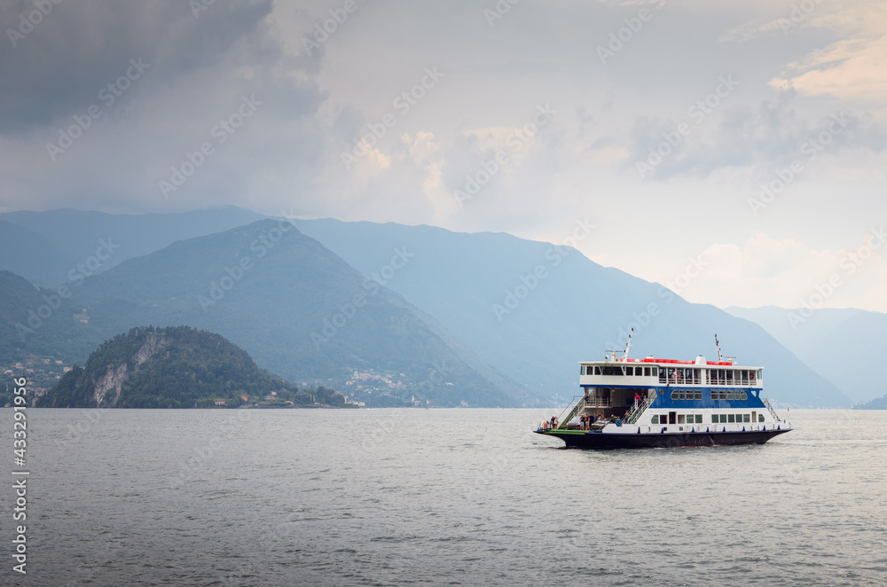 Early morning view of the Como Lake with a ferry boat sailing near the harbor of Varenna, Italy, cloudy sky, with fog, and mountains in the distance Early morning view of the Como Lake with a ferry boat sailing near the harbor of Varenna, Italy, cloudy sky, with fog, and mountains in the distance