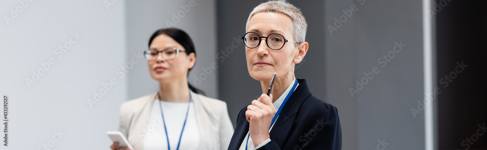 Mature businesswoman with pen standing near blurred asian colleague, banner Mature businesswoman with pen standing near blurred asian colleague, banner