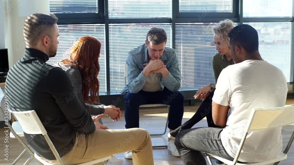 Front view of sad depressed young man telling about his mental problem or addiction to other patients sitting in circle. Multiethnic business people supporting upset male during group therapy session. Front view of sad depressed young man telling about his mental problem or addiction to other patients sitting in circle. Multiethnic business people supporting upset male during group therapy session.