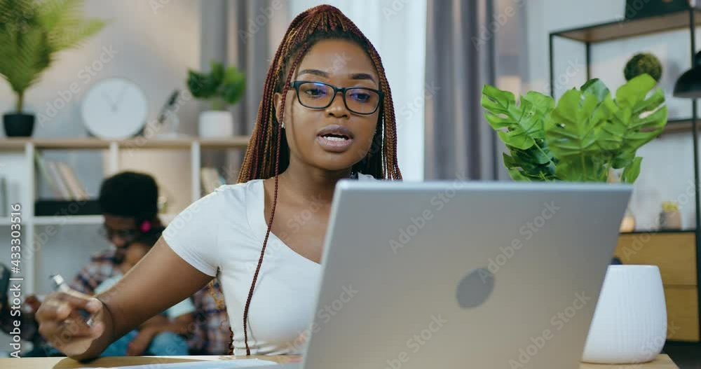 Focused african woman in eyewear working on wireless laptop at living room. Blur background of young man sitting on couch with kid and using smartphone.