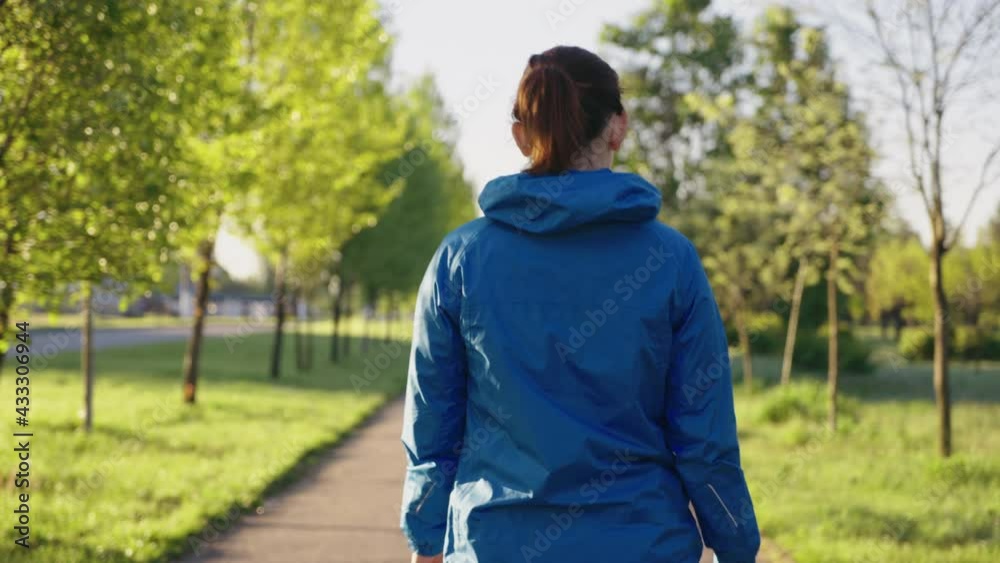 Woman wearing blue sports jacket walking in park at sunrise. Back view following shot female jogger after training outside. Concept of fitness Woman wearing blue sports jacket walking in park at sunrise. Back view following shot female jogger after training outside. Concept of fitness
