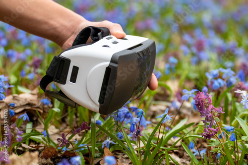 Hand holding 3D Virtual reality headset glasses in spring blue wild flowers. VR tech gear goggles close-up. Optical entertainment system for smartphones