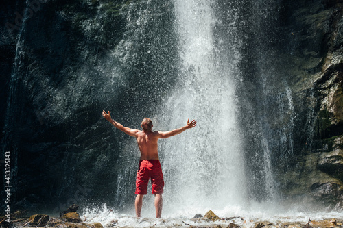 Middle-aged man dressed only red trekking shorts standing under the mountain river waterfall, rose arms up and enjoying the splashing Nature power. Traveling, trekking and nature concept image.