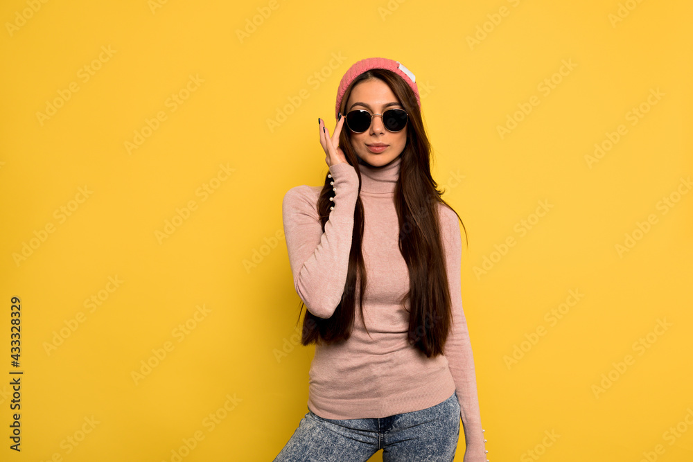 Fototapeta premium Elegant european girl with long dark hair wearing black sunglasses touching her glasses and posing before yellow wall