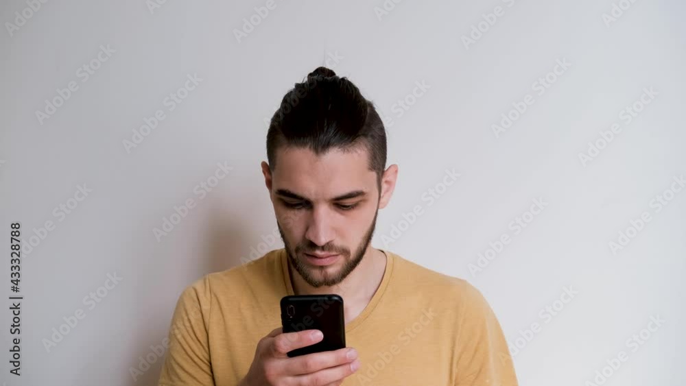 Young handsome man of Caucasian European appearance with beard and ponytail chats with mobile phone and looks carefully. Online correspondence using smartphone from home against white wall. Young handsome man of Caucasian European appearance with beard and ponytail chats with mobile phone and looks carefully. Online correspondence using smartphone from home against white wall.
