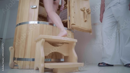 Girl with in swimsuit walks up an open cedar phyto barrel in spa salon close-up view. On the right is a master in a white work suit with slippers.