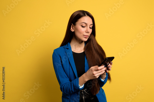 Charming lovely woman with long dark hair scrolling smartphone over yellow backdrop