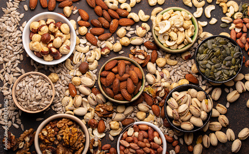 Selection of assorted raw nuts and various seeds in bowls on brown stone background from above, healthy source of energy, fat and vegetarian protein