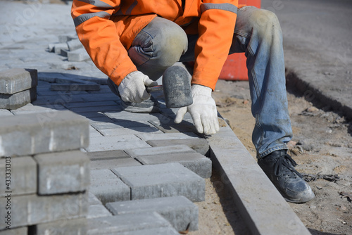 The worker knocks on the paving slabs with a construction hammer, aligns small concrete blocks laid in the sand, makes a pedestrian road.