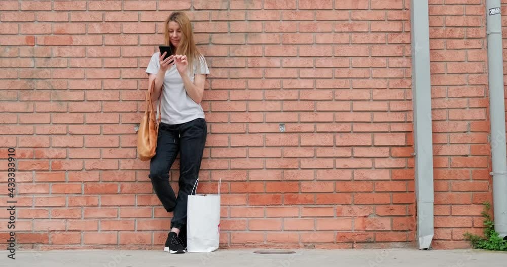 Young woman with shopping bag using smartphone while standing on the street by brick wall, concept taxi phone app, copy space. Young woman with shopping bag using smartphone while standing on the street by brick wall, concept taxi phone app, copy space.