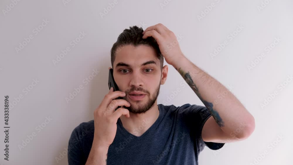 Young Caucasian man with beard and stylish hairstyle in blue Tshirt on white background is talking on phone, angry and waving his hands. Man throws smartphone aside after unpleasant conversation. Young Caucasian man with beard and stylish hairstyle in blue Tshirt on white background is talking on phone, angry and waving his hands. Man throws smartphone aside after unpleasant conversation.