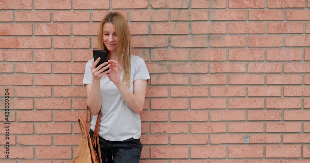 Young woman with shopping bag using smartphone while standing on the street by brick wall, concept taxi phone app, copy space. Young woman with shopping bag using smartphone while standing on the street by brick wall, concept taxi phone app, copy space.
