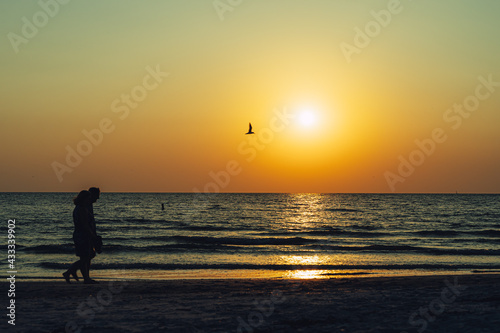 Two people walk along the beach in silhouette at sunset against orange sky
