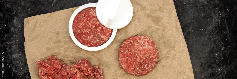 Patty Press with Burgers on Wooden Board on Black Background, Overhead View. Plastic Burger Press and Stuffer With Grill Tools. Beef and Pork Mince Patties. Patty Press with Burgers on Wooden Board on Black Background, Overhead View. Plastic Burger Press and Stuffer With Grill Tools. Beef and Pork Mince Patties.