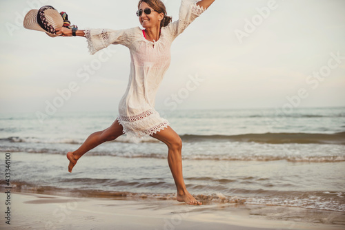 a woman with sunglasses and a hat in her hand jumps on the beach shore. she is happy. she is wearing a white thread dress. happiness and well-being. vacation at the seaside.