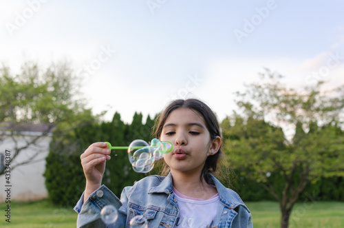 Hispanic girl blowing soap bubbles in the backyard with green grass.