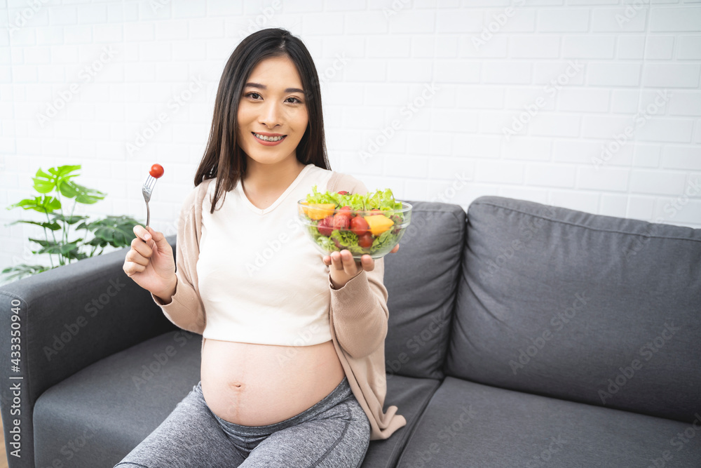 Beautiful asian pregnant woman holding a bowl of salad of vegetables and fruits, using a fork eating tomato. Eating healthy having lunch sitting on the grey sofa, relaxing and resting living room Beautiful asian pregnant woman holding a bowl of salad of vegetables and fruits, using a fork eating tomato. Eating healthy having lunch sitting on the grey sofa, relaxing and resting living room