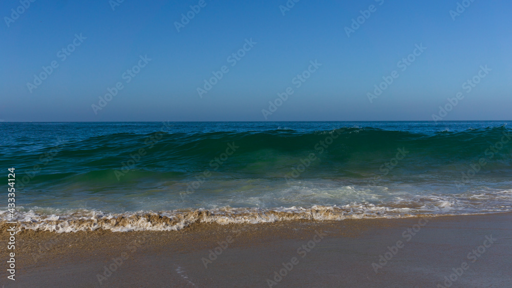 Wave about to crush onto sandy beach under blue sky