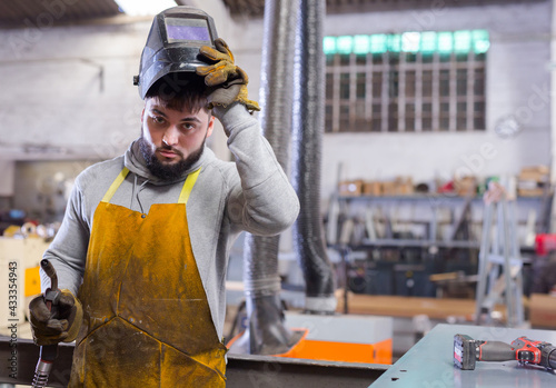 Portrait of young skilled welder wearing protective apron, gloves and helmet standing in metalworking workshop.. photo
