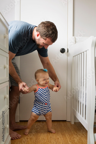 Mid adult man holding baby daughter hands whilst toddling in nursery