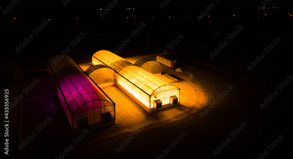 Drone view of two greenhouses implementing photosynthesis experiments with advanced agriculture are glowing in yellow and purple light during late evening.