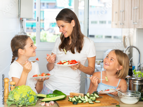 Young woman with two small girls enjoying salad at kitchen after cooking
