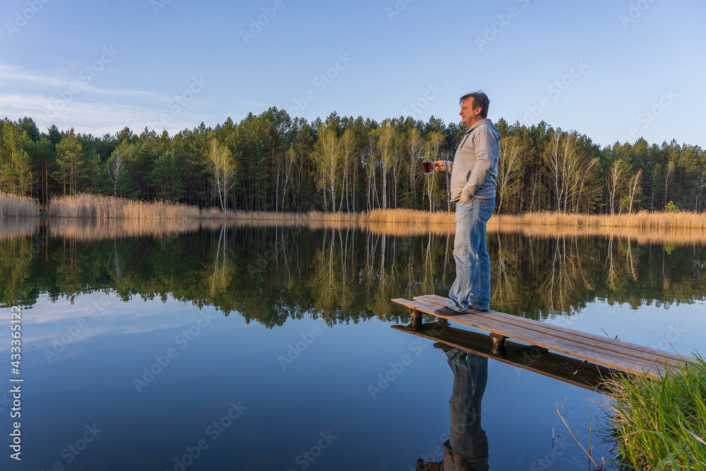 Happy man stands on a wooden pier with glass cup tea near spring forest on a calm lake. Nature and travel concept