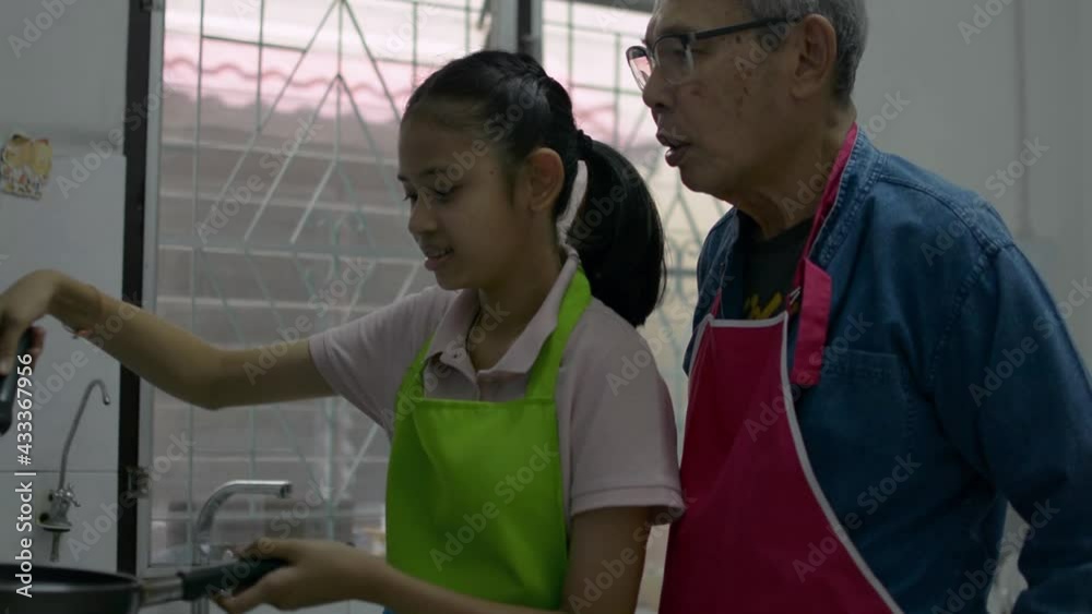 Happy Asian senior grandfather in apron teaching his adorable young granddaughter cooking food in a pan in the kitchen at home. Bonding relationship in family. Happy Asian senior grandfather in apron teaching his adorable young granddaughter cooking food in a pan in the kitchen at home. Bonding relationship in family.