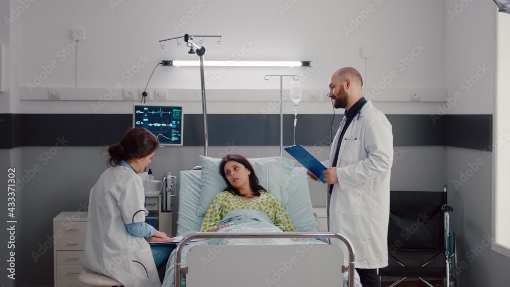 Specialist doctors monitoring sick woman discussing sickness treatment while working in hospital ward. Patient resting in bed explaining symptom disease during recovery appointment Specialist doctors monitoring sick woman discussing sickness treatment while working in hospital ward. Patient resting in bed explaining symptom disease during recovery appointment
