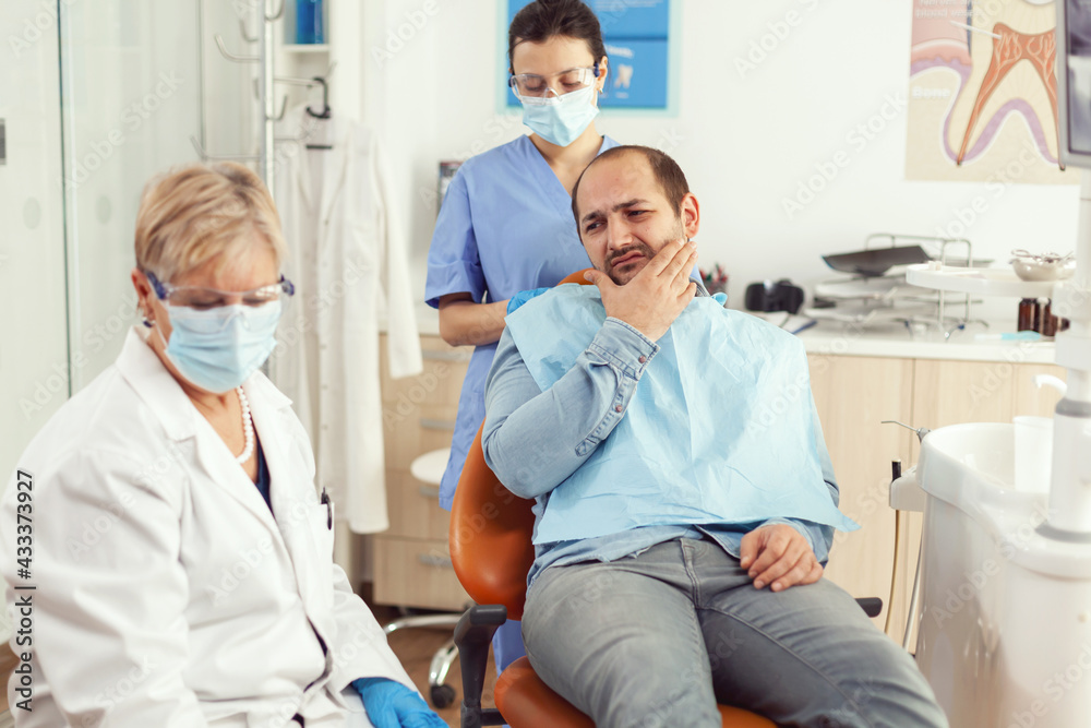 Man patient putting his hand on cheek showing toothache complaining about tooth pain. Senior woman explaining dental problem to doctor indicating mouth while sitting on stomatological chair Man patient putting his hand on cheek showing toothache complaining about tooth pain. Senior woman explaining dental problem to doctor indicating mouth while sitting on stomatological chair