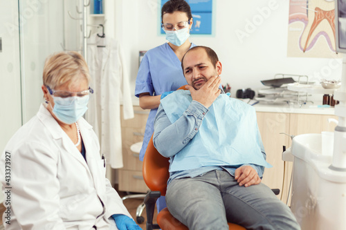 Man patient putting his hand on cheek showing toothache complaining about tooth pain. Senior woman explaining dental problem to doctor indicating mouth while sitting on stomatological chair