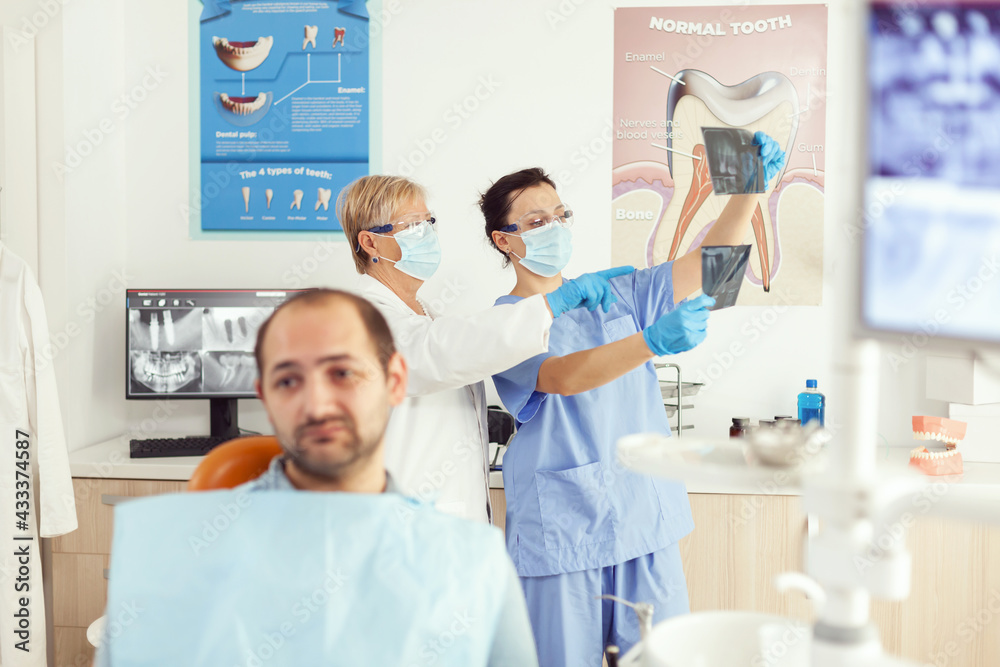 Obraz premium Sick man patient waiting for teeth examination sitting on dental chair in stomatology clinic room. Stomatologist doctor and medical nurse preparing for orthodontic surgery while examining radiography