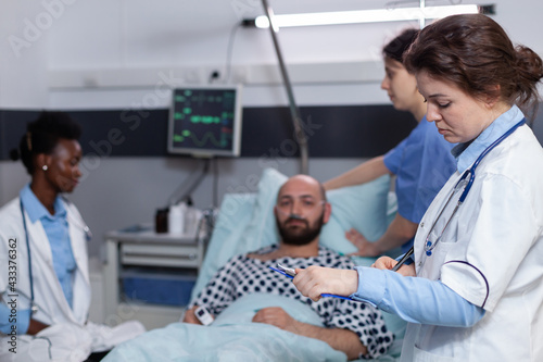 Front view of medical team monitoring sick man during recovery appointment in hospital ward. Specialist doctor writing disease treatment on clipboard examining patient symptom discussing sickness