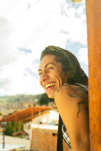 Retrato de mujer transgénero con trenzas riéndose  mientras sale a observar la ciudad desde la ventana de su habitación 