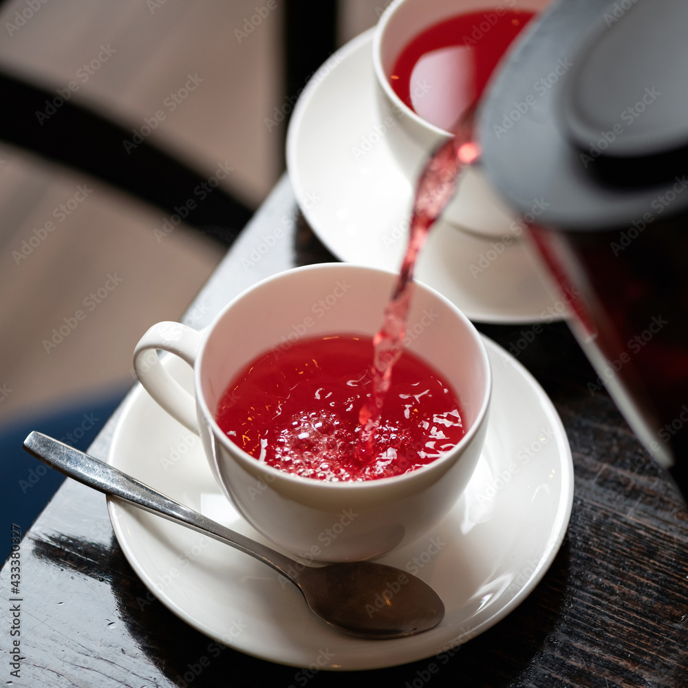 Red tea from frozen berries. Hot drink pouring from teapot into white cup and saucer on wooden table. Tea time. Close up shot. Soft focus. Red tea from frozen berries. Hot drink pouring from teapot into white cup and saucer on wooden table. Tea time. Close up shot. Soft focus.
