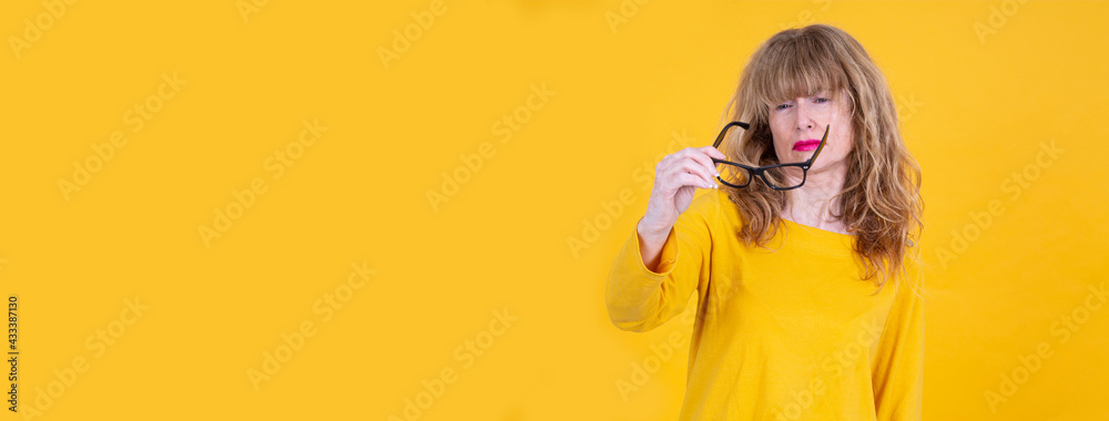 Woman has vision problems, squints her eyes while trying to see something, takes off her glasses, isolated on yellow background. Myopia, hyperopia, vision concept. See Less