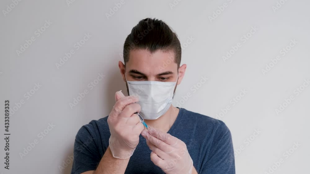 Young cute Caucasian student in blue Tshirt, medical protective mask and white latex gloves stands on white background and fills syringe with medicine. Man is going to give an injection. Young cute Caucasian student in blue Tshirt, medical protective mask and white latex gloves stands on white background and fills syringe with medicine. Man is going to give an injection.