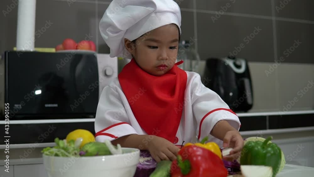 Cute asian girl wear chef uniform with a lot of vegatable on the table in the kitchen room,Make food for eat dinner,Funny time for kids Cute asian girl wear chef uniform with a lot of vegatable on the table in the kitchen room,Make food for eat dinner,Funny time for kids