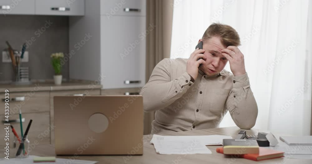 Man working at home sitting at his desk talking on the phone unpleasant conversation upset holding his head feels depressed crisis