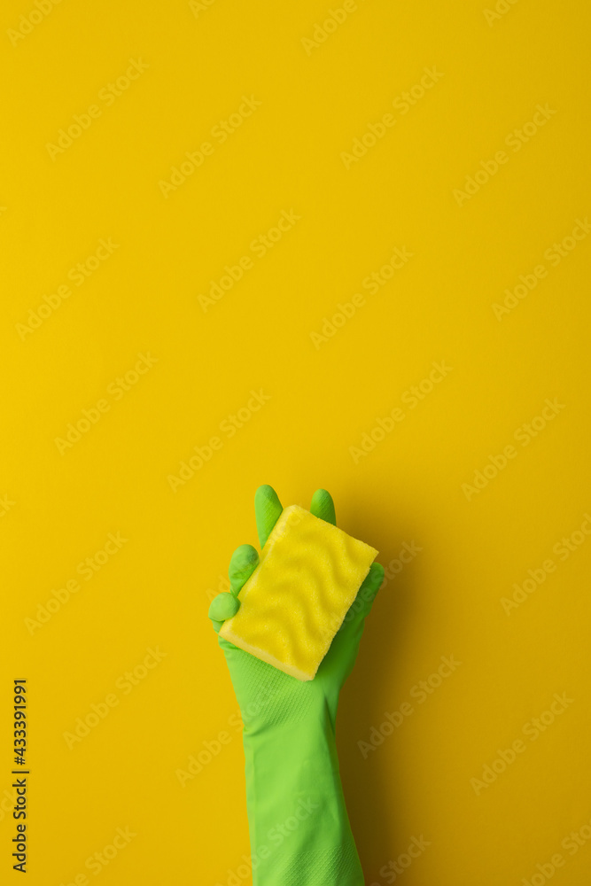 A hand in a green protective rubber glove holds a washing sponge on a yellow background A hand in a green protective rubber glove holds a washing sponge on a yellow background