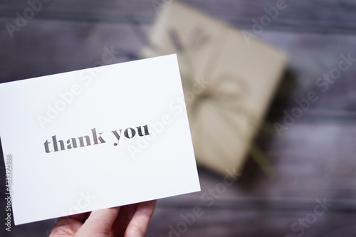 Woman's hand holding a thank you card with a present on wooden background. Special thank you note. Surprise gift. 