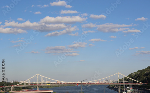 Beautiful steel bridge over the river on a sunny day. Scenic morning blue sky with little fluffy clouds. People walking outdoors. © Liudmyla Leshchynets
