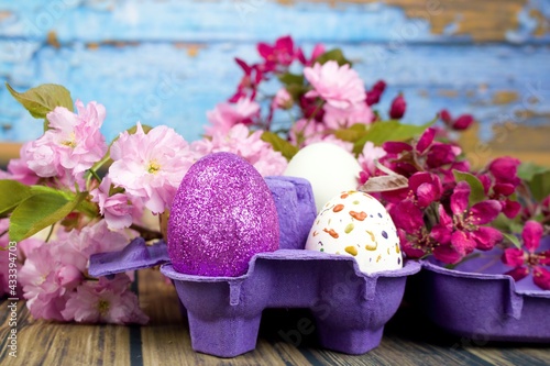 Four eggs decorated with wax and glitter in a purple egg wrapper and branches of ornamental cherries.