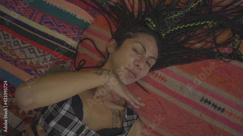 Mujer transgénero con trenzas y tatuajes haciendo gestos acostada sobre la alfombra de su habitación 