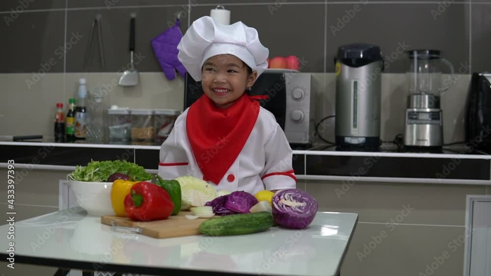 Cute asian girl wear chef uniform with a lot of vegatable on the table in the kitchen room,Make food for eat dinner,Funny time for kids Cute asian girl wear chef uniform with a lot of vegatable on the table in the kitchen room,Make food for eat dinner,Funny time for kids