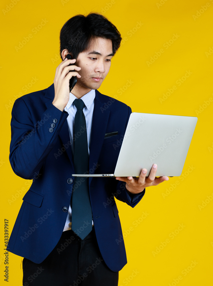 Young Asian businessman in formal blue suit. Standing work with laptop. Other hand holding phone seems for a call. Yellow background gives modern feel. The space can be filled with text. Young Asian businessman in formal blue suit. Standing work with laptop. Other hand holding phone seems for a call. Yellow background gives modern feel. The space can be filled with text.