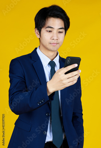 Handsome Businessman in formal blue suit. Standing look at mobile phone seems texting message. Yellow background gives modern feel. The space can fill with text.