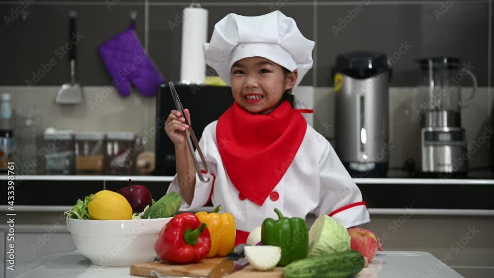 Cute asian girl wear chef uniform with a lot of vegatable on the table in the kitchen room,Make food for eat dinner,Funny time for kids Cute asian girl wear chef uniform with a lot of vegatable on the table in the kitchen room,Make food for eat dinner,Funny time for kids