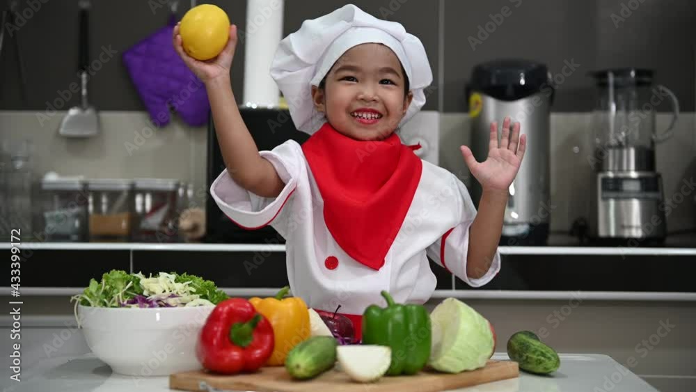 Cute asian girl wear chef uniform with a lot of vegatable on the table in the kitchen room,Make food for eat dinner,Funny time for kids Cute asian girl wear chef uniform with a lot of vegatable on the table in the kitchen room,Make food for eat dinner,Funny time for kids