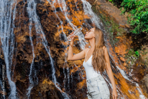 A young woman drinks clean water from a bottle in the jungle with a waterfall in the background. Healthy clean natural water. 